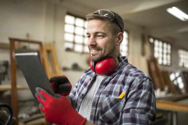 Lächelnder junger Handwerker/Ingenieur arbeitet in der Werkstatt lächelnd auf seinem Tablet mit Handschuhen.