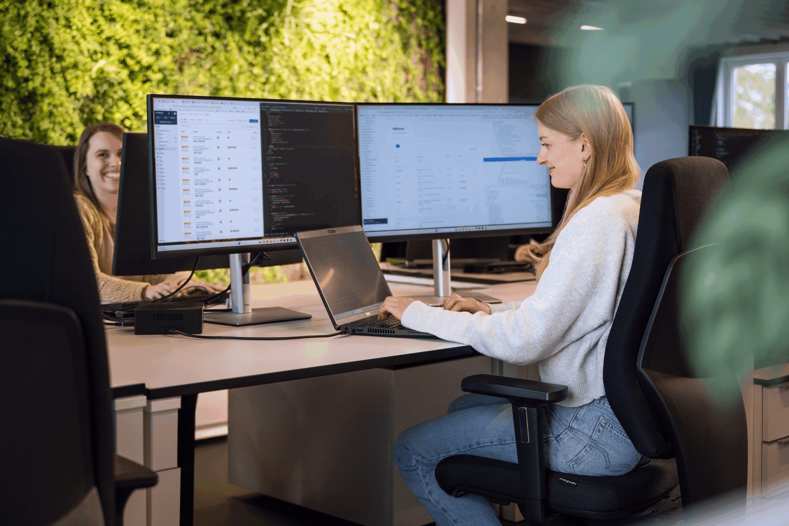 Mitarbeiterin von Worksdone arbeitet im modernen Büro an einem Laptop und mehreren Bildschirmen; im Hintergrund sitzt eine Kollegin vor einer begrünten Wand.